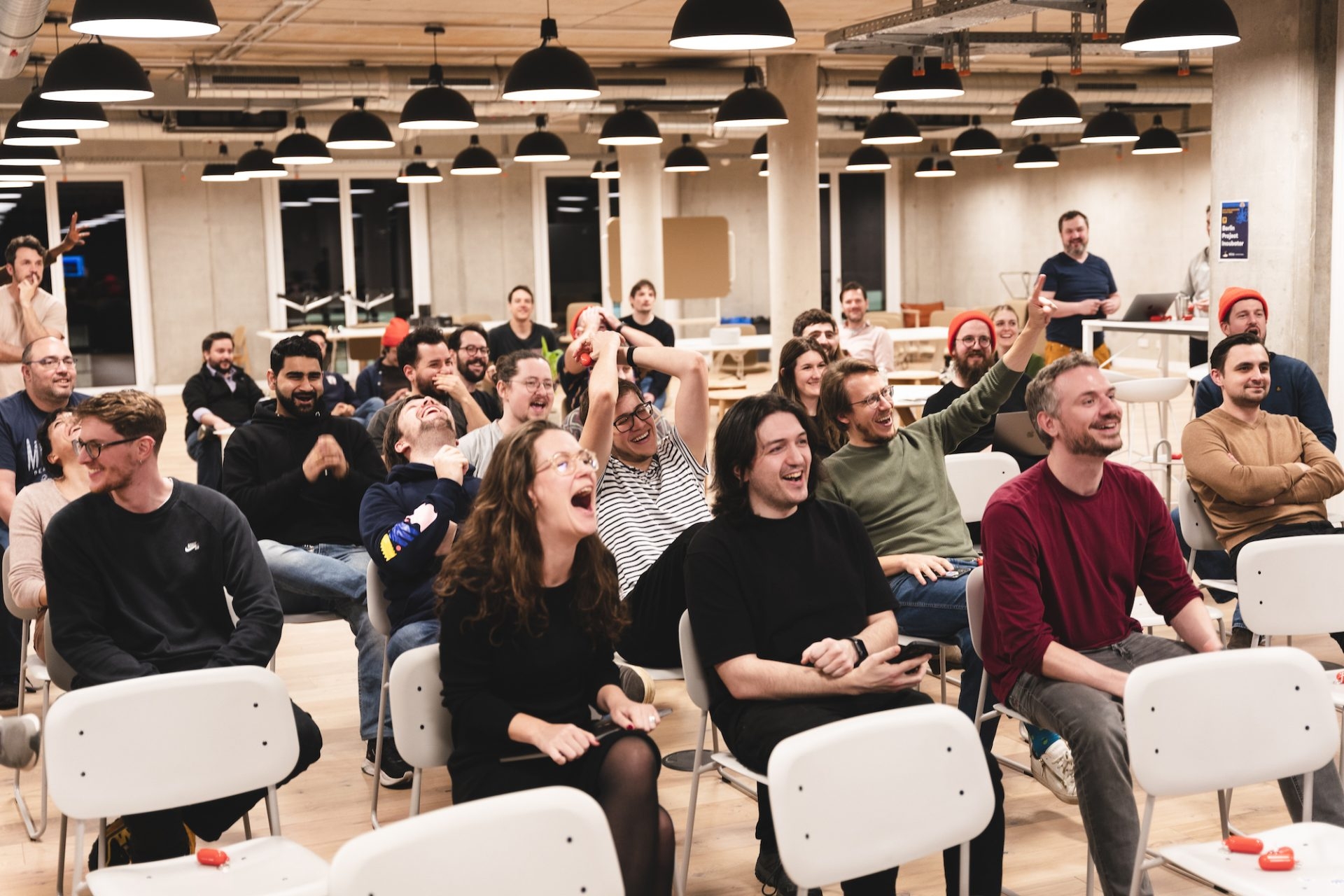 A diverse group of Miro employees sit in chairs arranged in an open-plan office, laughing and enjoying a presentation.