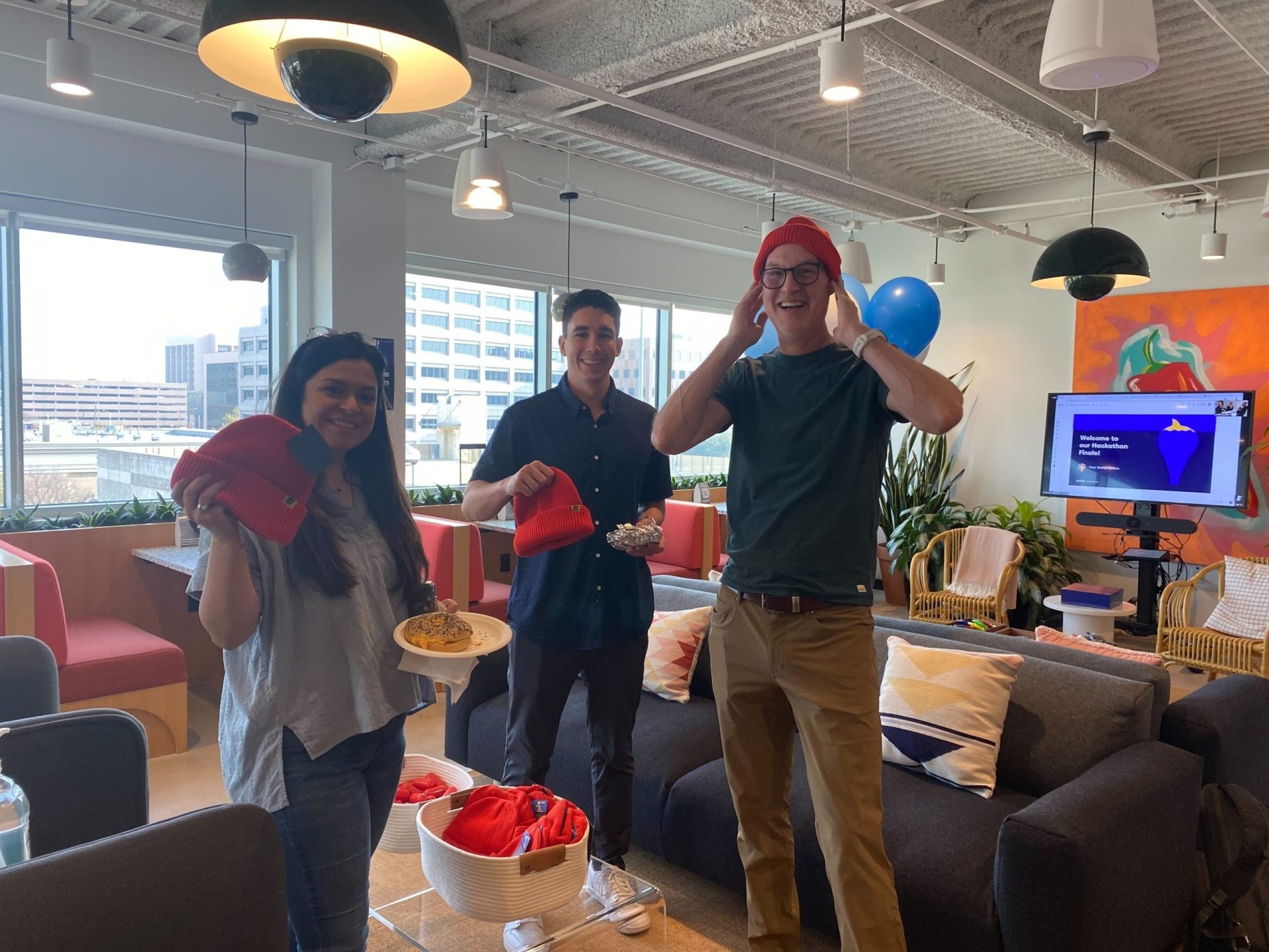 Three Miro employees smile at the camera in a hang-out spot in an office. One, a tall man, wears an orange beanie. The other two, a man and a woman, smile and hold up their own orange beanies.